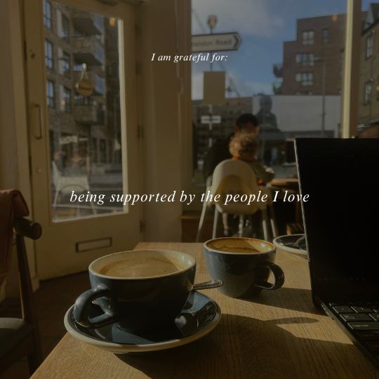 Two coffee cups on a table in a coffee shop in a sunny winter day. The text on the photo says: I am grateful for being supported by the people I love.